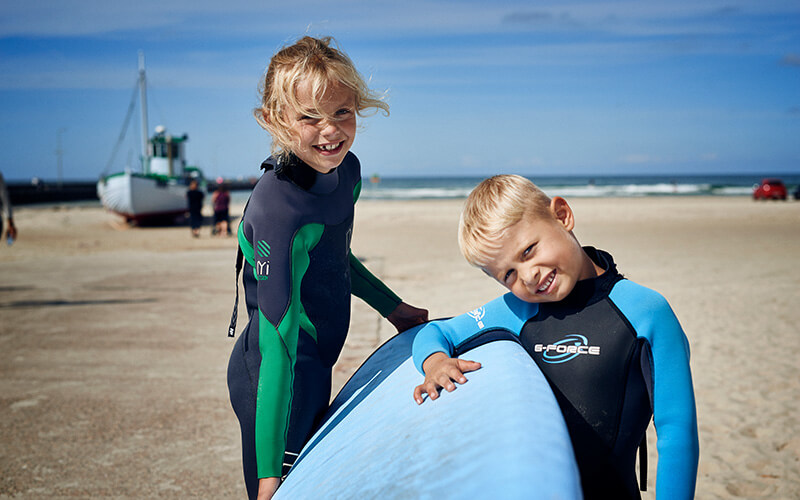 Zwei Kinder in Neoprenanzügen stehen am Sandstrand und halten ein blaues Surfboard.