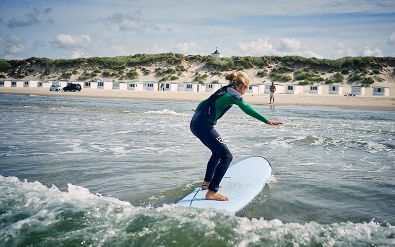 Surfer:in im Neoprenanzug steht auf einem Surfbrett in Wellen nahe einem Strand mit Dünen und Strandhäusern im Hintergrund.