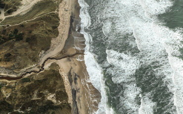 Blick von oben auf Meer und Strand mit Schaumkronen auf dem Wasser und Pflanzen hinter dem Strand.