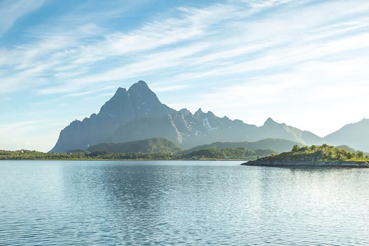 Bergkette mit markanten Gipfeln hinter ruhigem Fjordwasser unter leicht bewölktem Himmel.