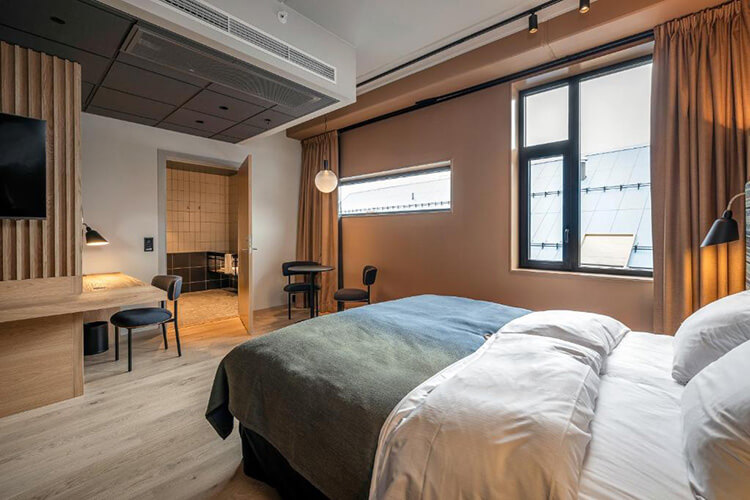 Modern eingerichtetes Hotelzimmer mit großem Bett, Schreibtisch, Stühlen und Fenster mit Blick auf ein Dach.