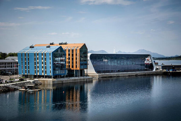 Moderne Hotelgebäude mit großen Glasfronten am Wasser, im Hintergrund Berge unter bewölktem Himmel.