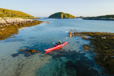 Zwei Personen paddeln in roten Kajaks auf klarem Wasser zwischen felsigen Uferbereichen und kleinen Inseln.