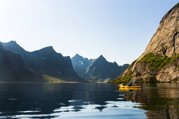 Zwei Personen paddeln in gelben Kajaks auf ruhigem Wasser vor steilen, felsigen Bergen unter klarem Himmel.