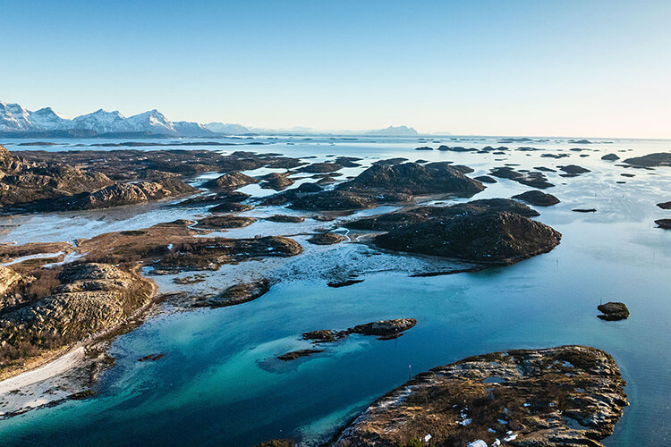 Luftaufnahme einer zerklüfteten Fjordlandschaft mit kleinen Inseln und schneebedeckten Bergen im Hintergrund.