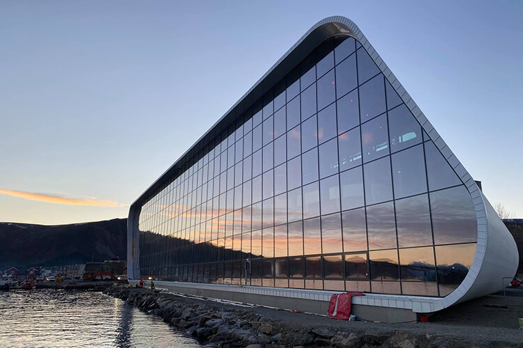 Modernes, langgestrecktes Gebäude mit großer Glasfassade, das Hurtigsten Museum in Stokmarknes, das sich an einem Fjordufer befindet, im Abendlicht.