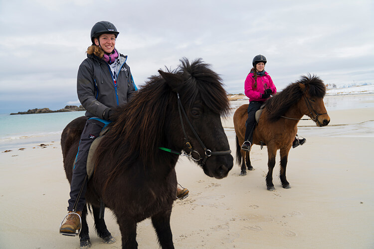 Zwei Personen reiten auf Islandpferden am Strand, beide tragen Reithelme und wetterfeste Kleidung.