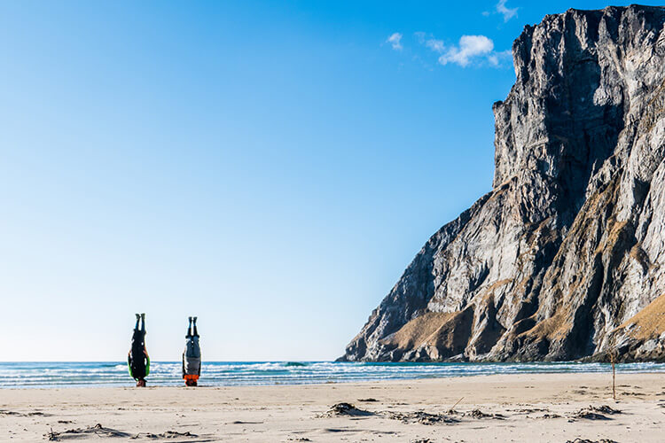 Zwei Personen stehen auf dem Kopf am Strand vor einem hohen Felsen unter blauem Himmel.