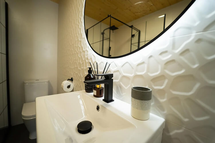 Weißes Waschbecken mit schwarzem Wasserhahn und Duftstäbchen vor einer Wand und rundem Spiegel, im Hintergrund Toilette und Duschkabine.