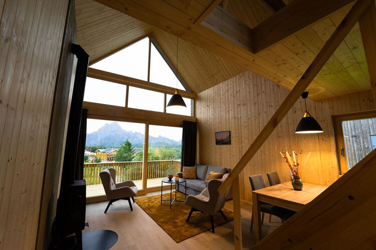 Innenraum einer Holzhütte mit großen Fenstern, zwei Sesseln, Sofa, Esstisch mit Stühlen und Blick auf Berge.