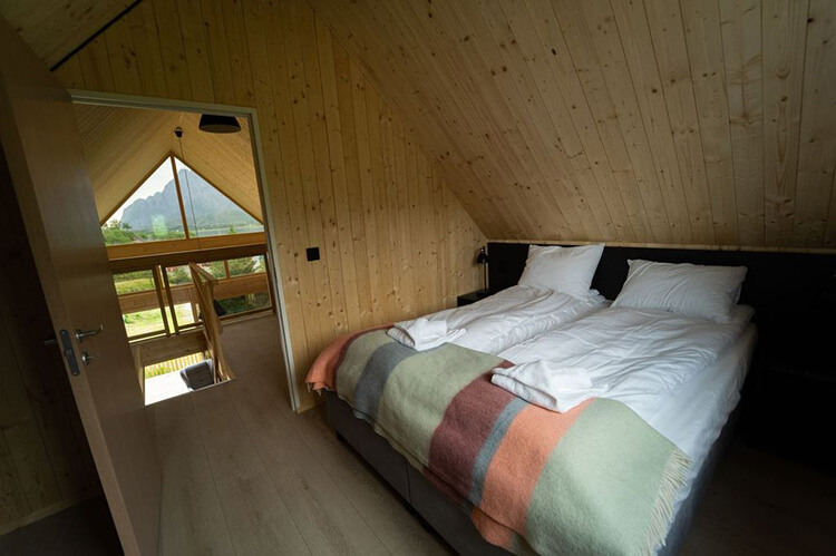 Schlafzimmer mit Doppelbett, Holzwänden und Blick durch Fenster auf eine Terrasse und Berge.