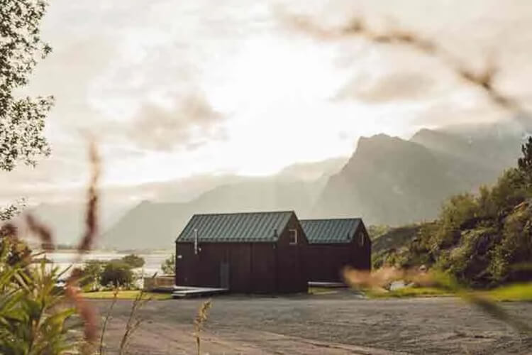 Zwei kleine Holzhäuser mit Metall-Dächern vor einem Fjord und Bergen im Hintergrund bei bewölktem Himmel.