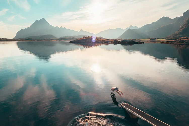 Person springt von einem Steg in einen ruhigen Fjord, umgeben von Bergen unter bewölktem Himmel.
