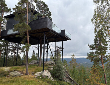 Modernes Baumhaus auf Stelzen in bewaldeter, hügeliger Landschaft mit Glasgeländer und Blick ins Tal.