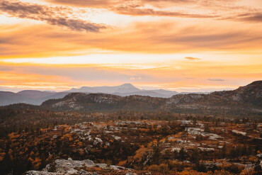 Weite Berglandschaft mit Felsen und vereinzeltem Bewuchs unter einem orangefarbenen Himmel bei Sonnenuntergang.