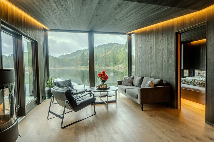 Wohnzimmer mit großen Fenstern, zwei Sesseln, Sofa, runden Tisch mit Blumen und Blick auf bewaldeten See.