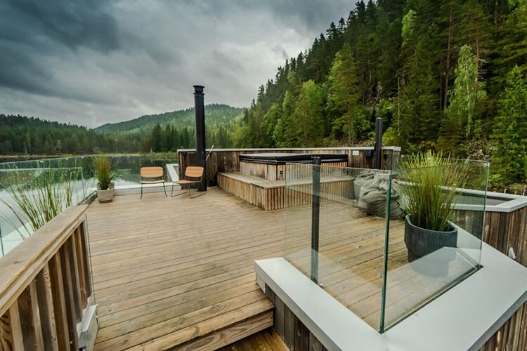 Holzterrasse mit Whirlpool, Sitzgelegenheiten und Glasgeländer vor bewaldeten Hügeln unter bewölktem Himmel.