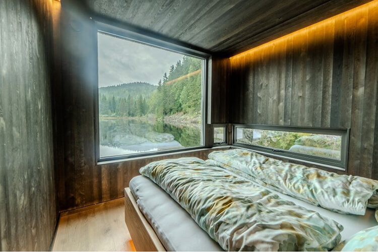 Schlafzimmer mit großem Bett und zwei großen Fenstern, die auf einen bewaldeten See blicken.