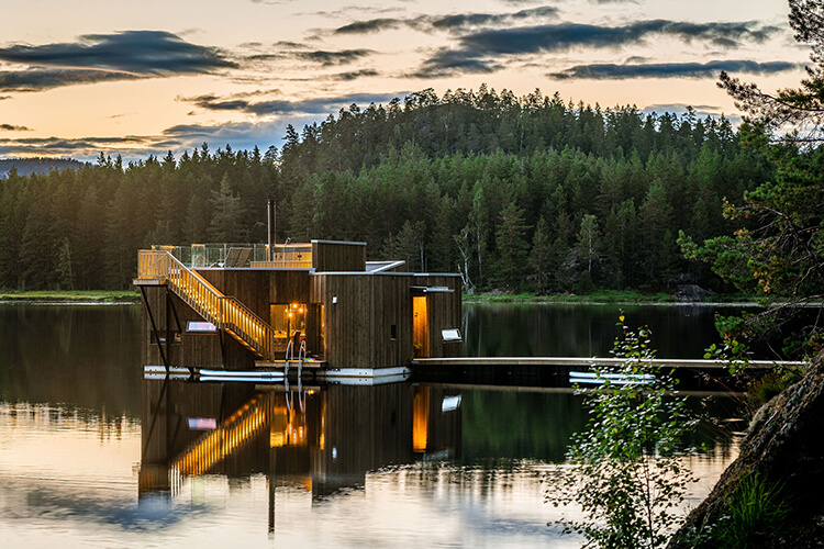 Modernes, beleuchtetes Holzhaus auf einem See, umgeben von Wald und bewölktem Himmel bei Dämmerung.