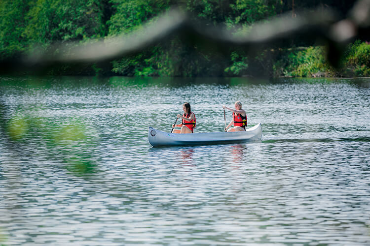 Zwei Personen mit roten Schwimmwesten paddeln in einem weißen Kanu auf einem ruhigen See, umgeben von grünem Wald.