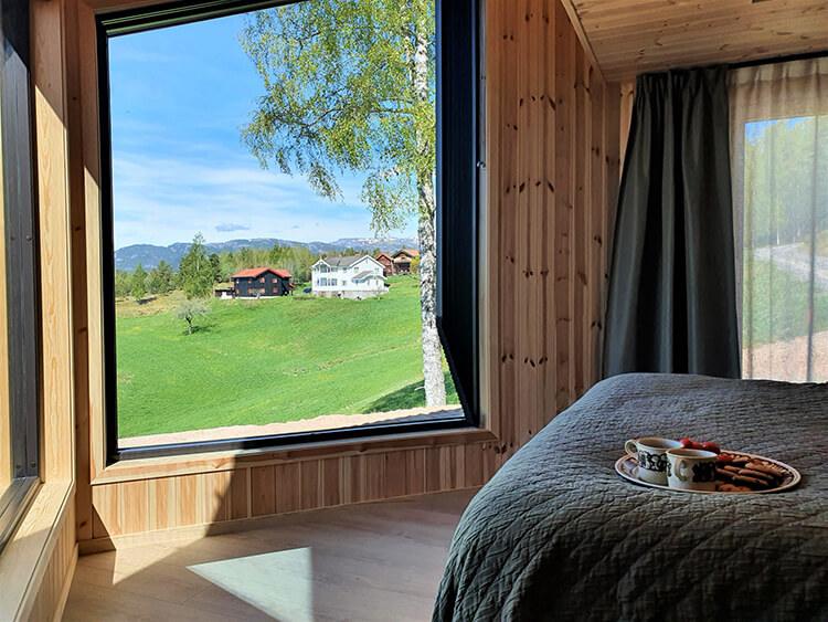 Blick aus einem Holzhausfenster auf grüne Wiese, Bäume und mehrere Häuser vor Bergen, im Vordergrund Bett mit Tablett und Tasse.