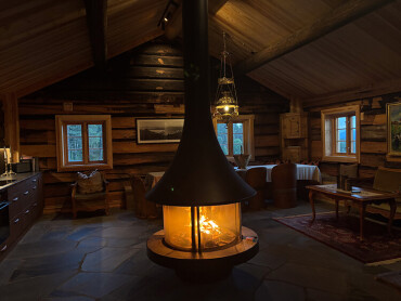 Offener Kamin mit brennendem Feuer in einer rustikalen Holzhütte mit Holzmöbeln und Fenstern.