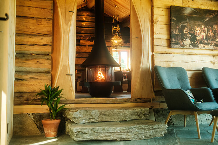 Offener Kamin mit Feuer in einer rustikalen Holzhütte, daneben ein Topfpflanze und ein blauer Sessel.