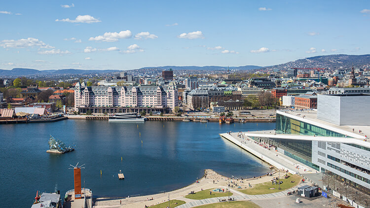 Blick auf den Oslofjord mit dem markanten weißen Opernhaus am Ufer und historischen Gebäuden im Hintergrund, umgeben von Stadt und Bergen.