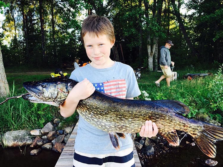 Kind steht auf einem Steg am Ufer und hält einen großen Hecht mit beiden Händen