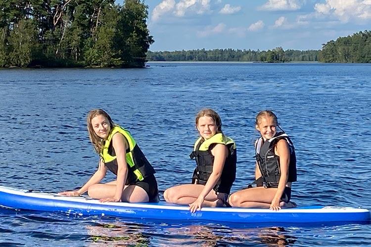 Drei Personen mit Schwimmwesten sitzen auf einem großen Stand-up-Paddleboard auf einem See mit bewaldetem Ufer im Hintergrund.