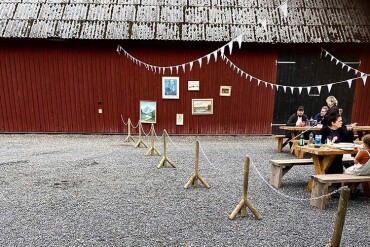 Mehrere Personen sitzen an Holzbänken vor einer rot gestrichenen Scheunenwand mit mehreren gerahmten Bildern und einer weißen Wimpelkette darüber