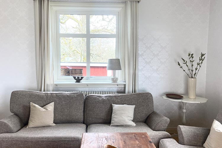 Wohnzimmer mit grauem Sofa, zwei Kissen, einem Sessel, einem Holztisch, Fenster mit weißen Vorhängen und einem kleinen Beistelltisch mit Vase und Zweigen