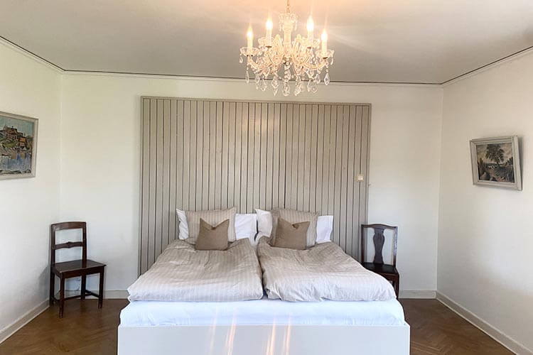 Schlicht eingerichtetes Schlafzimmer mit Doppelbett, zwei Stühlen an den Seiten und zwei gerahmten Bildern an den weißen Wänden