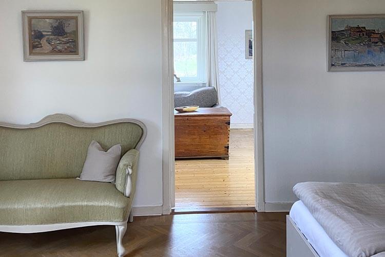 Blick durch eine offene Tür von einem Raum mit grünem Sofa und Holzboden in einen weiteren Raum mit Holzboden, Fenster und Holzkiste