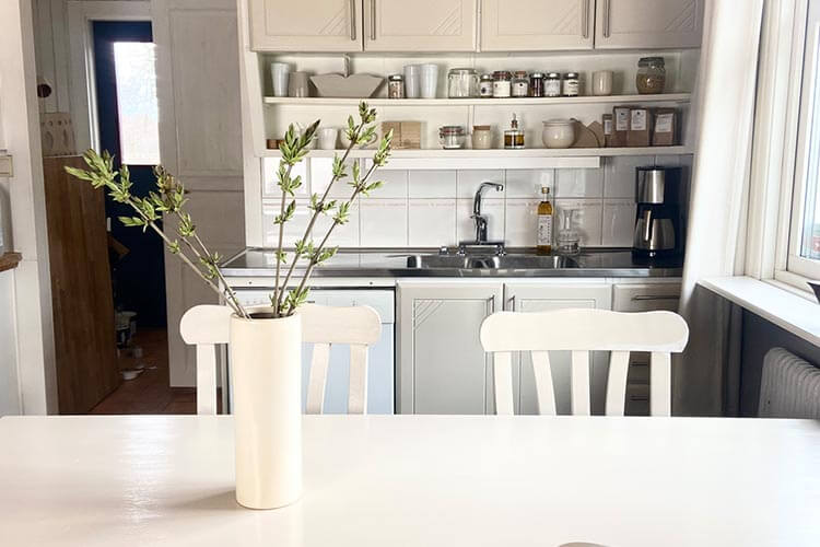 Helle Landhausküche mit weißem Tisch, zwei weißen Stühlen und Vase mit Zweigen auf dem Tisch, im Hintergrund Spüle, Regale mit Gläsern und Gewürzen sowie Kaffeemaschine