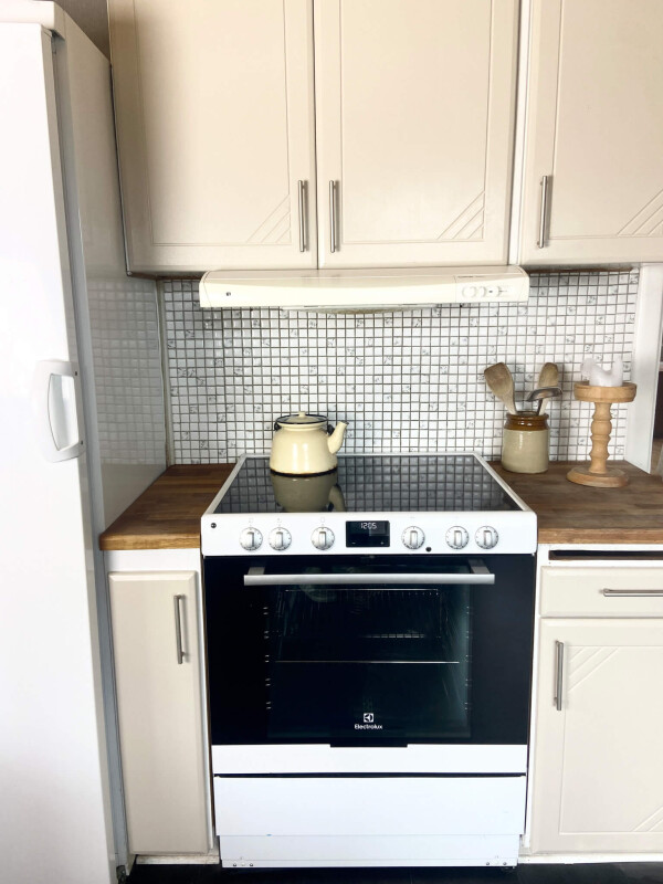 Küchenecke mit weißem Elektroherd, weißem Kühlschrank links, Holzarbeitsplatten und weißen Oberschränken vor einer Wand mit kleinen quadratischen Fliesen.