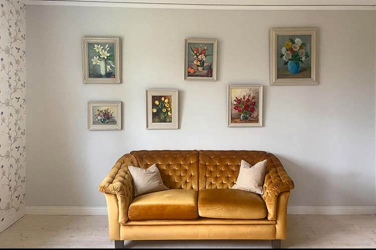 Gelbes Samtsofa mit zwei hellen Kissen vor weißer Wand mit sechs gerahmten Blumenbildern in verschiedenen Größen