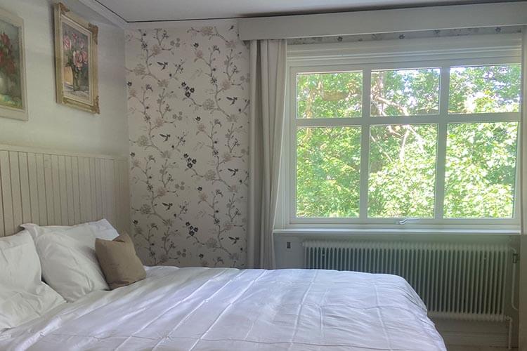 Schlafzimmer mit großem Bett, floral gemusterter Tapete an einer Wand, zwei gerahmten Bildern und einem Fenster mit Blick auf grüne Bäume