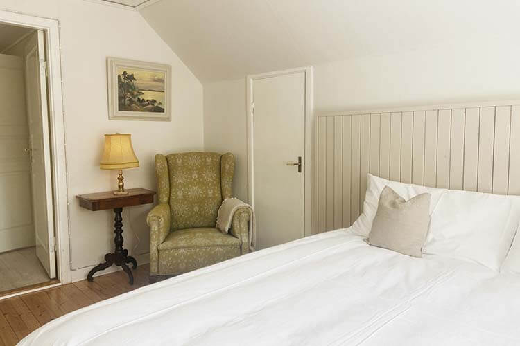 Schlafzimmer mit weiß bezogenem Bett, grün gemustertem Sessel, Nachttisch mit Lampe und einem Bild an der Wand