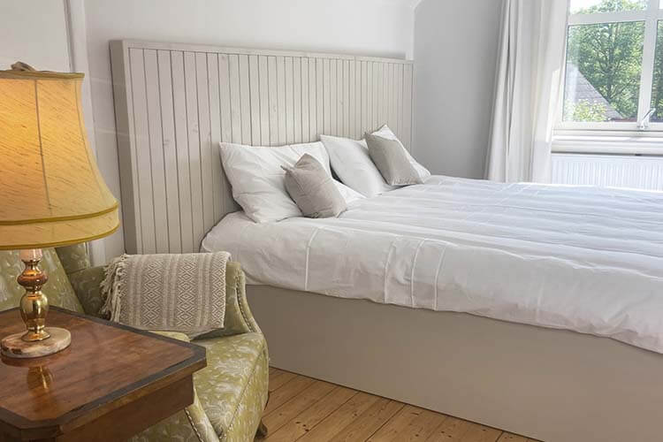 Schlafzimmer mit weiß bezogenem Bett, grün gemustertem Sessel, Nachttisch mit Lampe und einem Bild an der Wand