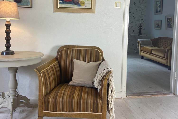 Gestreifter Sessel mit Kissen und Decke neben einem runden Beistelltisch mit Lampe in einem hellen Raum mit Blick in ein weiteres Zimmer mit Sofa.