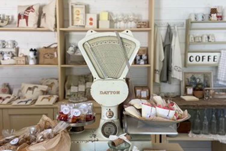 Antike Waage steht auf einem Ladentisch vor Regalen mit Kissen, Gläsern und weiteren Haushaltswaren.