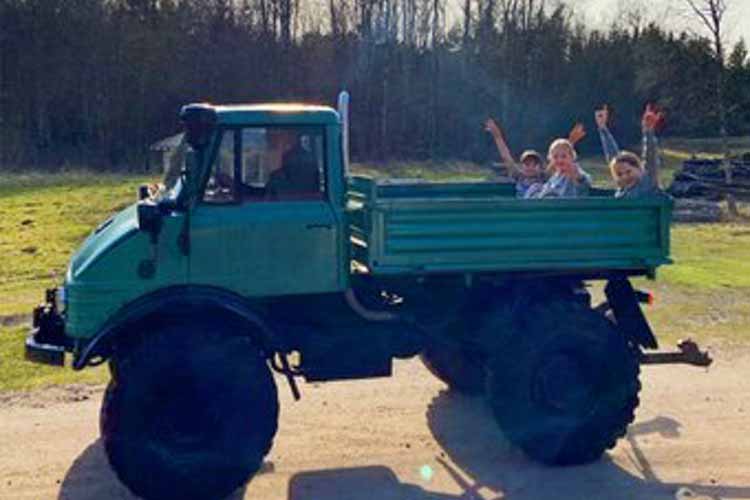 Vier Personen sitzen im offenen Ladebereich eines grünen Unimog-Geländefahrzeugs auf einem Feldweg vor einem Wald