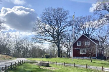 Rotes Bauernhaus mit weißem Fensterrahmen und Fahnenmast auf grünem Grundstück mit kahlen Bäumen und blauem Himmel mit Wolken