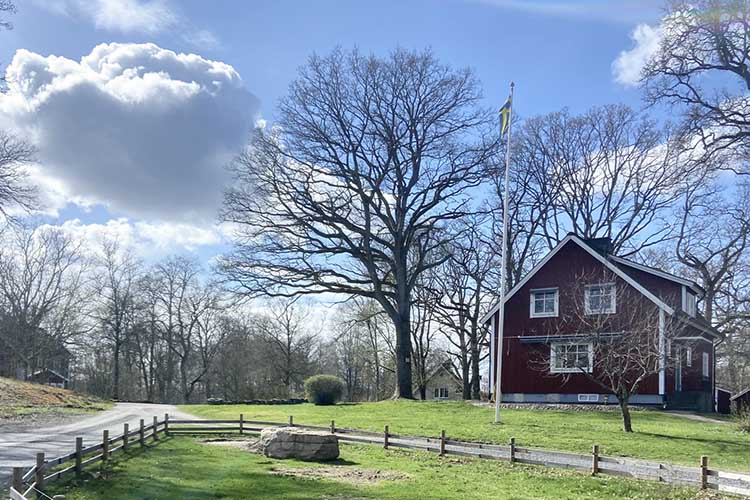 Rotes Bauernhaus mit weißem Fensterrahmen und Fahnenmast auf grünem Grundstück mit kahlen Bäumen und blauem Himmel mit Wolken