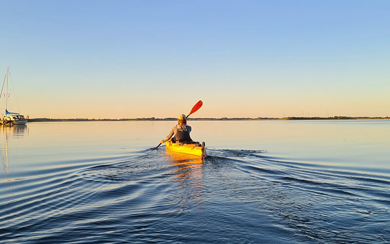Person in gelbem Kajak paddelt auf ruhigem Gewässer mit Segelboot am linken Bildrand.
