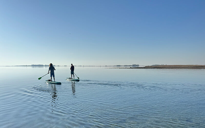 Zwei Personen stehen auf Stand-up-Paddle-Boards auf ruhigem Wasser unter klarem Himmel.