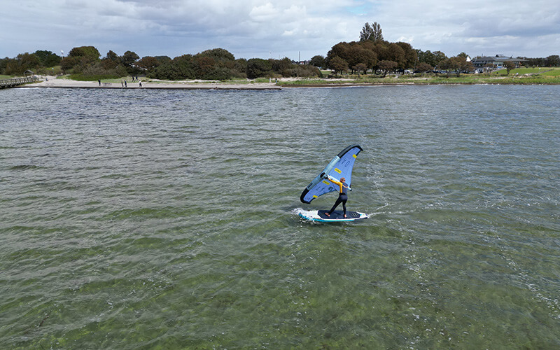 Windsurfer auf grünlichem Wasser vor flachem Ufer mit Strand, Bäumen und Menschen.