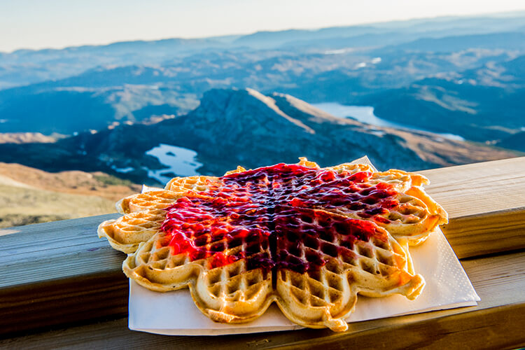 Eine Waffel mit Marmelade liegt auf einem Geländer, im Hintergrund sieht man eine Berglandschaft.