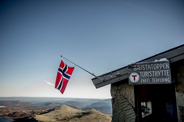 Norwegische Flagge an einer Berghütte mit Schild 'GAUSTATOPPEN TURISTHYTTE ENKEL SERVERING' vor weitem Bergpanorama